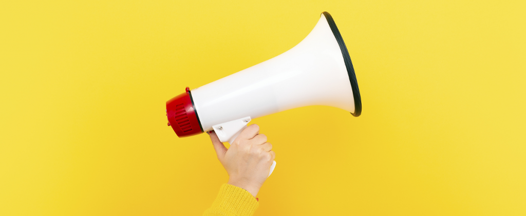 A hand holding up a white megaphone in front of a bright yellow wall