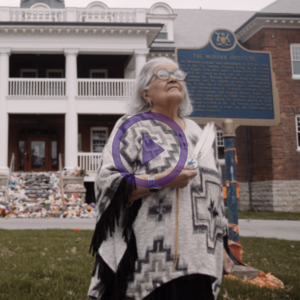 Elder Jan Longboat standing in front of the Mohawk Institute Residential School holding a feather and looking up into the sky