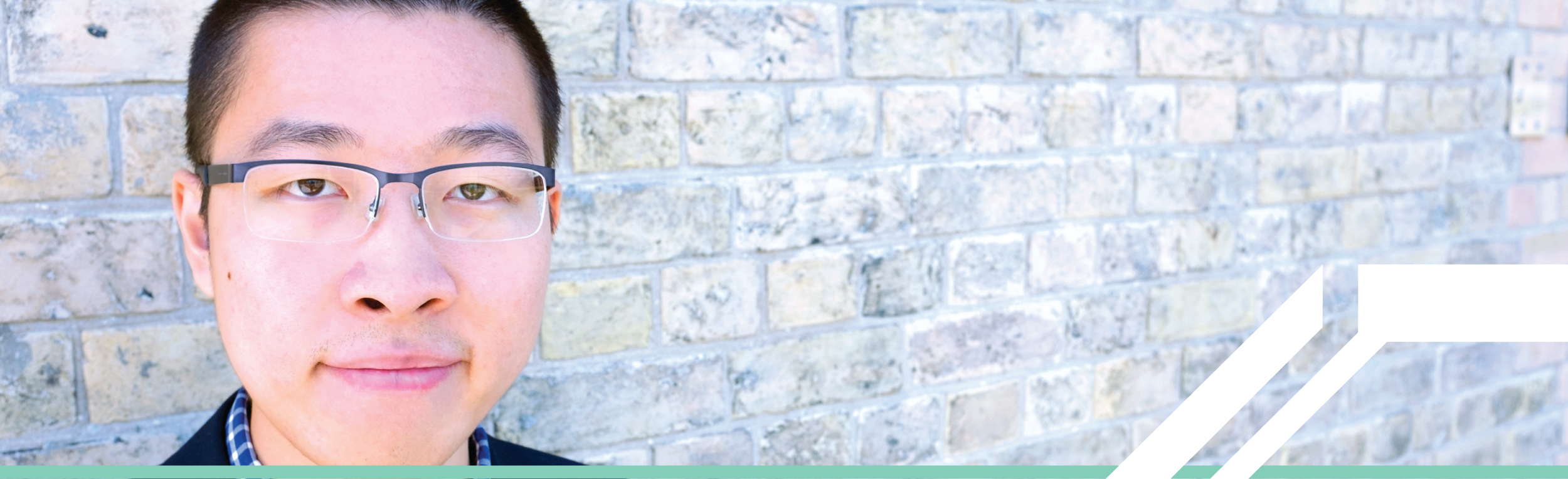 An Asian man with short dark hair, wearing glasses, standing in front of a grey brick wall