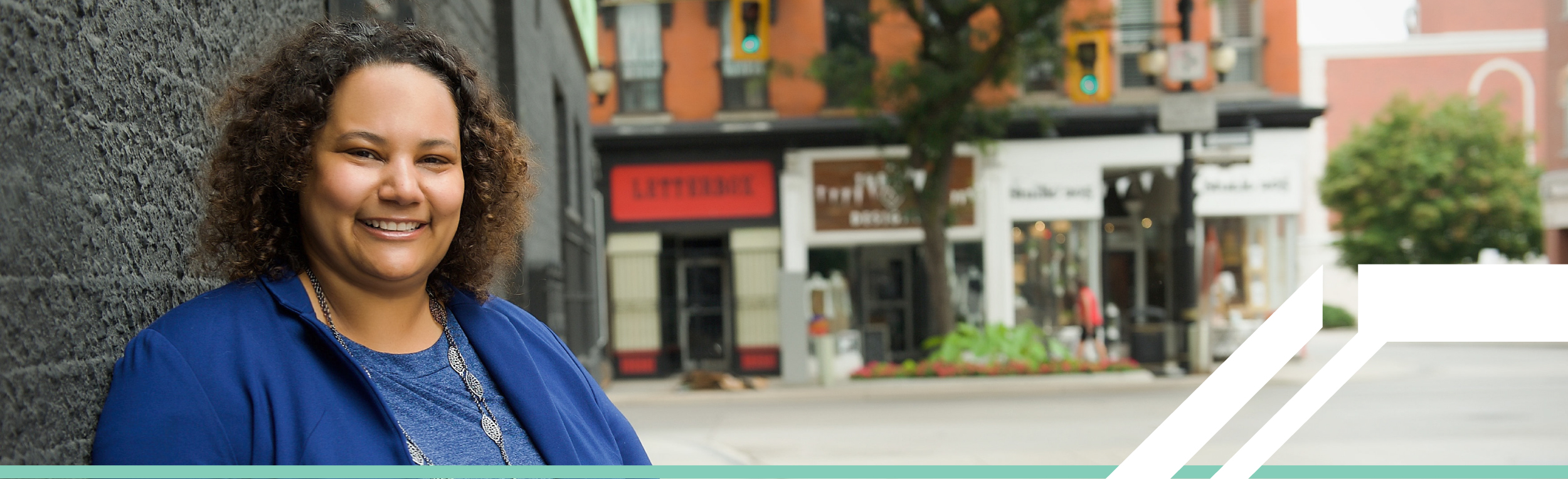 Une femme noire, souriante, portant une chemise et une veste bleue roi, se tient à l'extérieur d'une rue calme de la ville