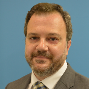 Man with brown hair and beard smiling softly into camera, wearing white dress shirt and grey suit jacket