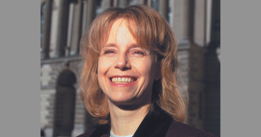 Woman smiling widely, who has shoulder-length blonde hair and standing in front of old buildings on University of Toronto campus