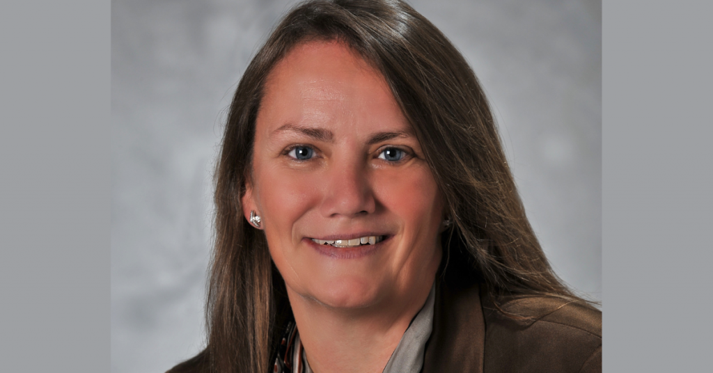 Woman with long dark hair, smiling and wearing small diamond and silver earrings and a brown jacket