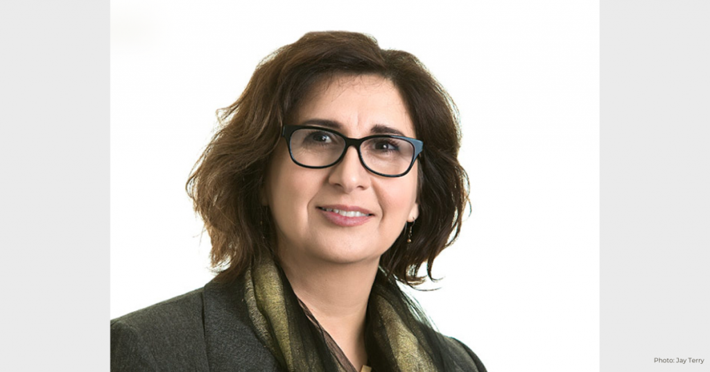 Woman with shoulder-length dark hair, wearing dark glasses and a light brown scarf and jacket