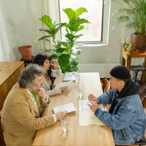 Three people sit at a wooden table in a bright, plant-filled room