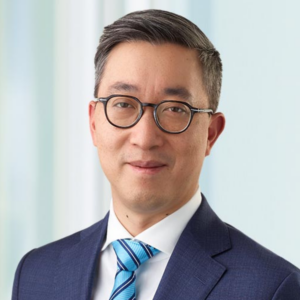 Man smiling slightly who has short black hair and dark glasses and is wearing a light and dark blue tie