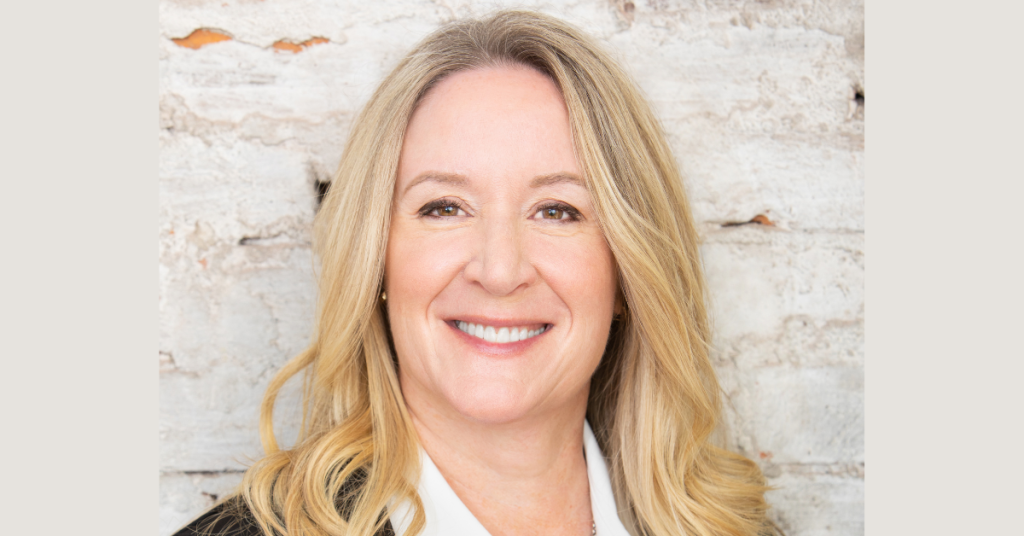 A woman with long blonde hair, smiling and wearing a white shirt and black blazer