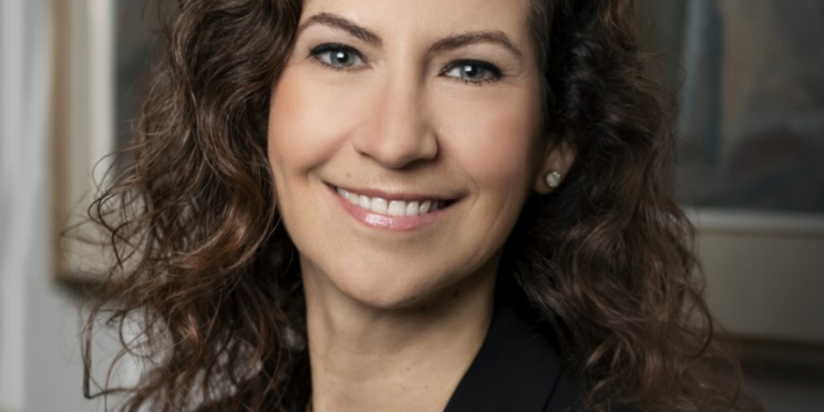 Woman with long, dark brown curly hair. She is smiling and wearing a black suit jacket.