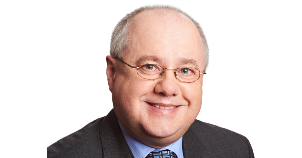 A man, smiling, who has very short grey hair and round silver-coloured glasses