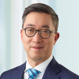 Man with short black hair and dark glasses who is smiling slightly and is wearing a navy suit jacket and a tie that is light and dark blue stripes