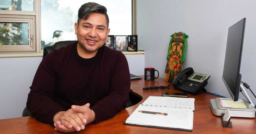 A man with salt and pepper hair, sitting at his office desk, and smiling.