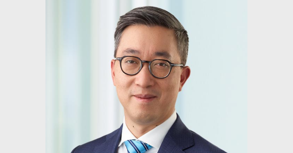 Man with short black hair and dark glasses who is smiling slightly and is wearing a navy suit jacket and a tie that is light and dark blue stripes