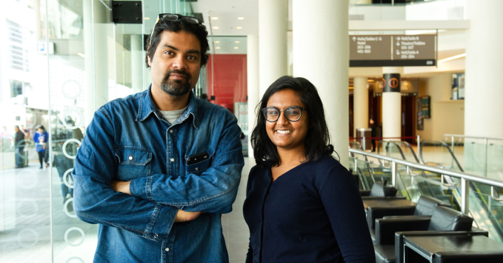 A man and woman standing beside each other, smiling. The man has dark hair and a moustache and goatee and is wearing a blue denim shirt. The woman has shoulder length black hair and is wearing dark framed glasses and a dark blue shirt.