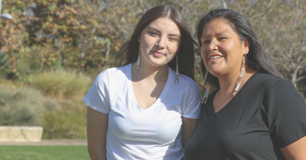 A young Indigenous woman standing with an older Indigenous woman, both smiling