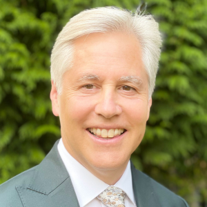 A man with white hair who is smiling and wearing a grey suit jacket. He is standing outside in front of thick, vivid green bushes