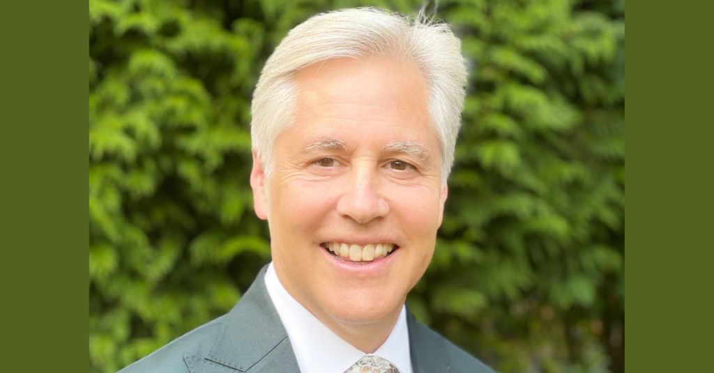 A man with white hair who is smiling and wearing a grey suit jacket. He is standing outside in front of thick, vivid green bushes