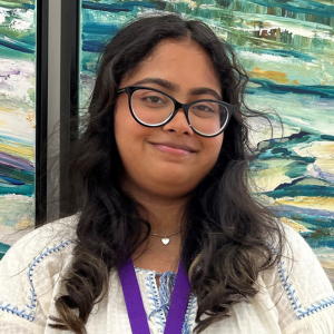 Young woman with long dark curly hair and glasses. She is smiling slightly and wearing a white shirt with a colourful painting in the background.