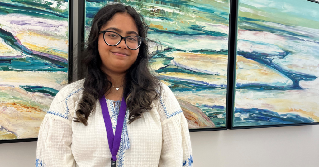 Young woman with long dark curly hair and glasses. She is smiling slightly and wearing a white shirt with a colourful painting in the background.