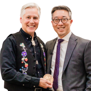 Two men smiling. The man on the left has white hair and is holding an award he received.