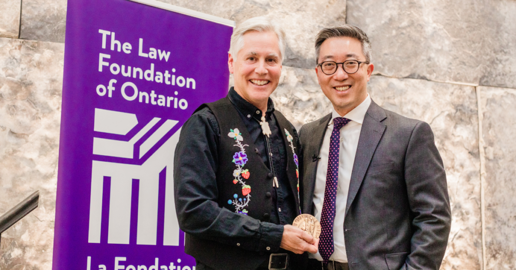 Two men standing beside each other smiling. The man on the left has white hair and a black shirt with Indigenous cultural beading and he is holding an award he received. The man on the right has dark hair and glasses.