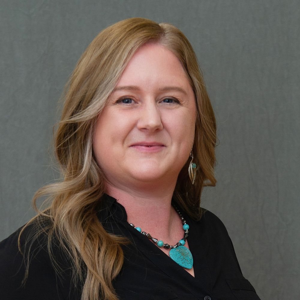 Woman with long blond wavy hair wearing a black jacket and teal necklace