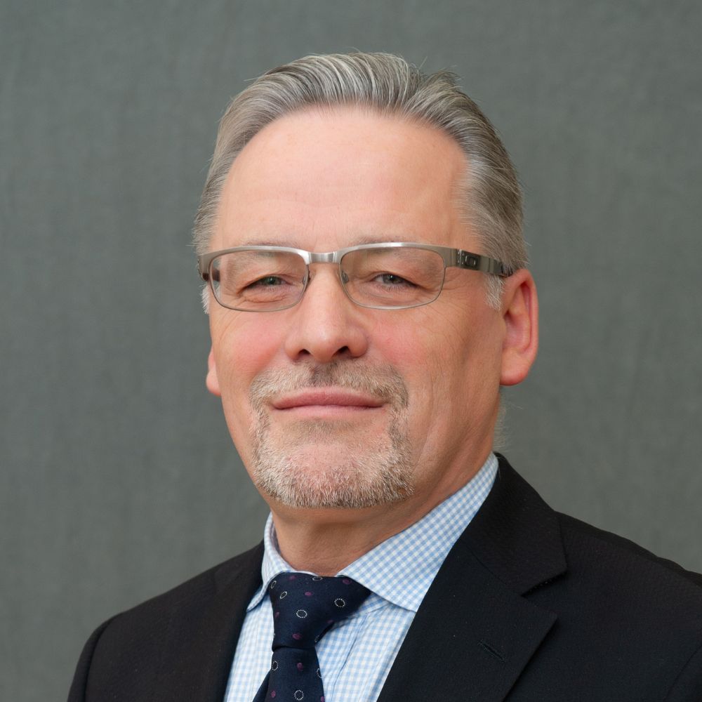 A man with grey hair and goatee wearing silver glasses, a dark coloured tie and suit jacket and light blue checkered shirt