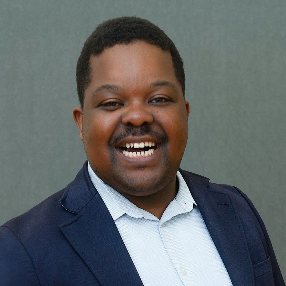 Man with very short black hair wearing a navy blue jacket and light blue shirt