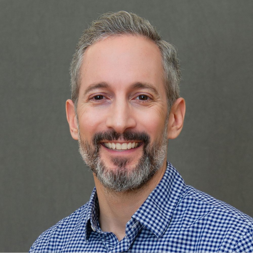 Man with short grey hair and beard wearing a blue and white checkered shirt