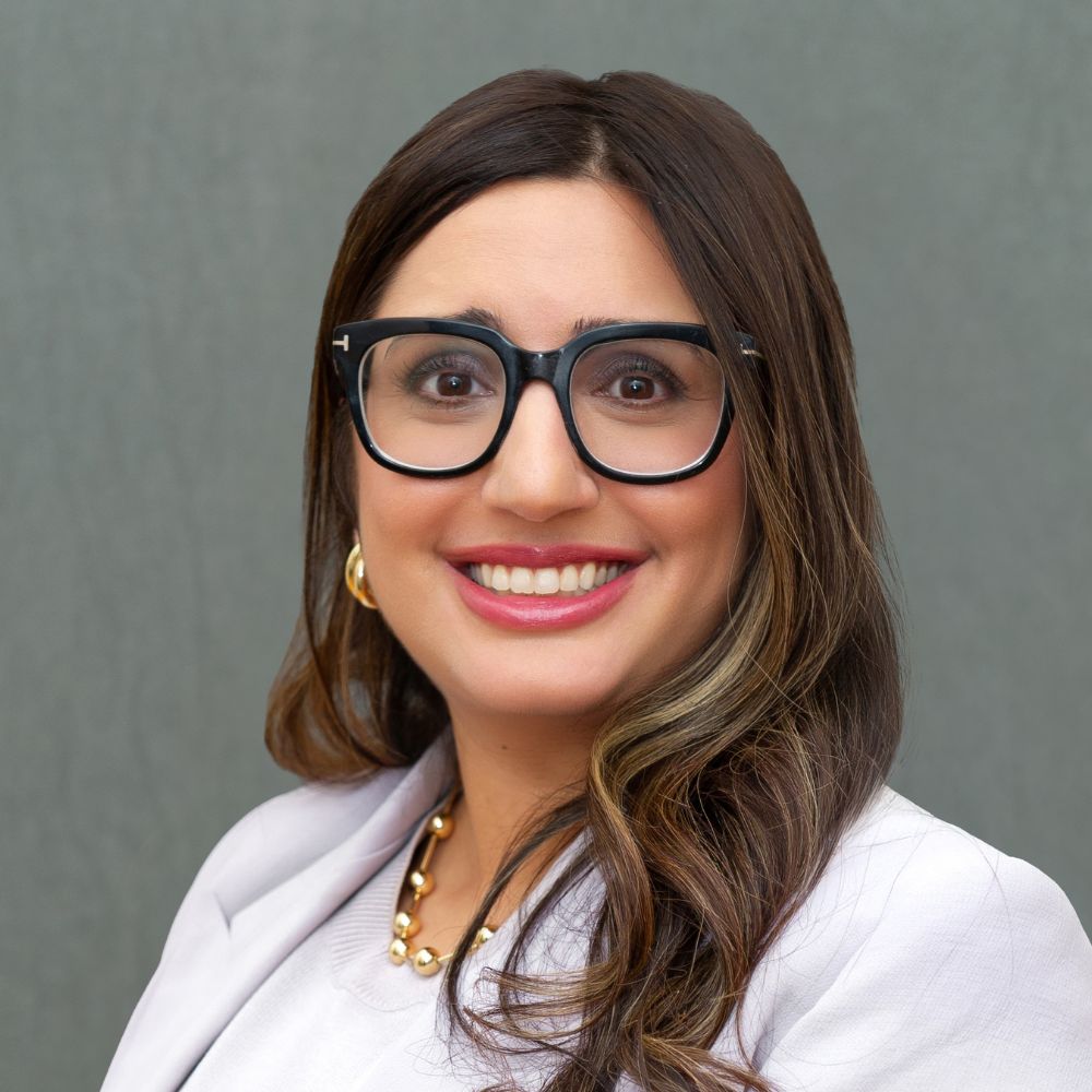 Woman with long dark brown hair wearing black glasses and a lilac-coloured suit jacket and shirt