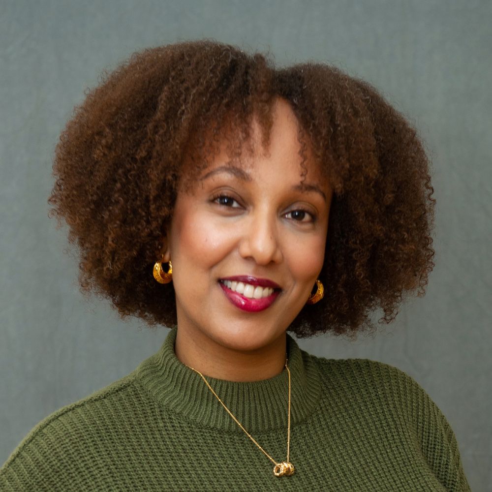 Woman with very curly dark brown hair wearing a green top and small gold necklace