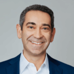 A man with short black hair who is smiling and wearing a white dress shirt and dark blue blazer