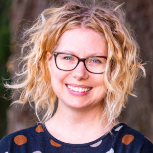 A woman with chin-length curly blonde hair wearing glasses and a polka dot shirt